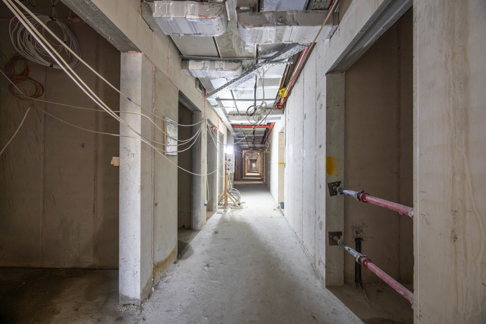 Cove Edition I By Imtiaz 1 A long, empty corridor in a building under construction, featuring exposed pipes and electrical wiring.
